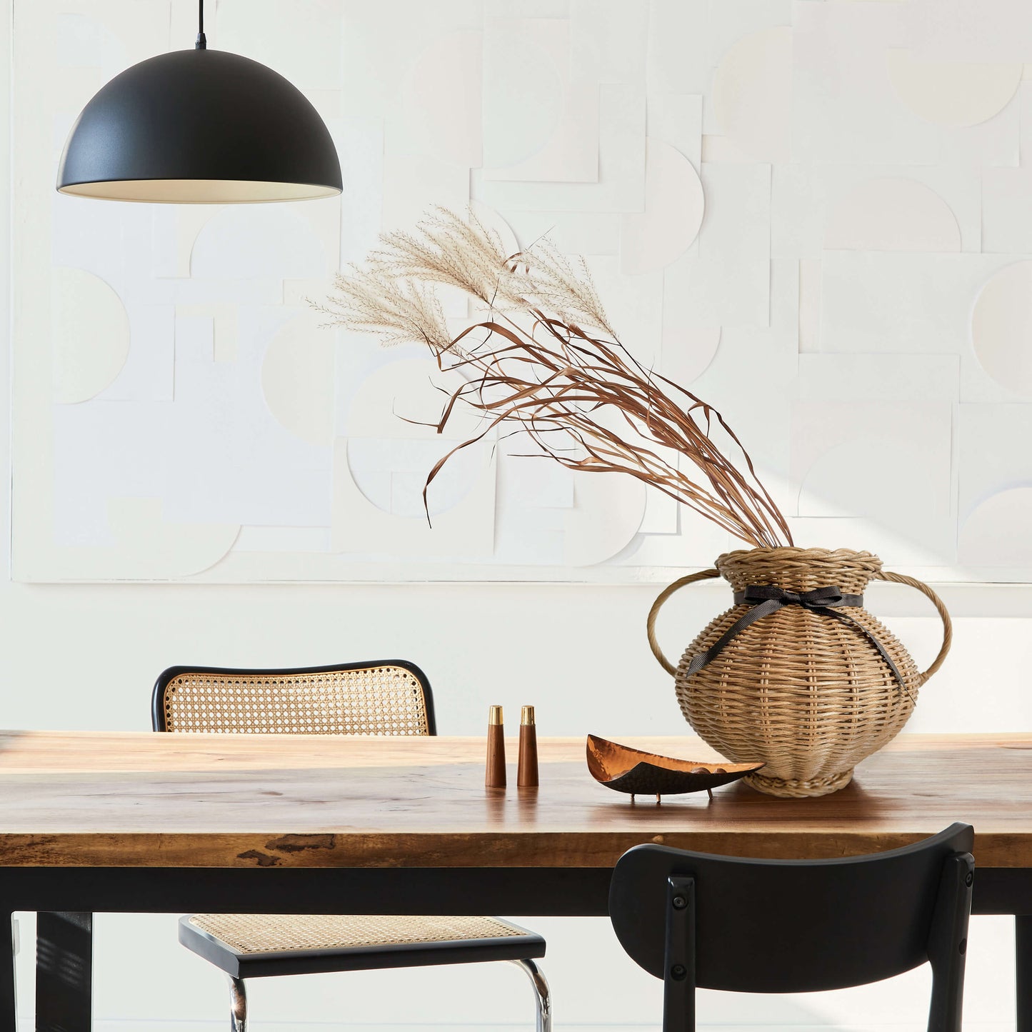 Kulplu el yapımı rattan vazo, siyah kurdele ile bağlanmış, yayvan formlu. Modern ahşap yemek masası üzerinde, kuru otlarla dekore edilmiş